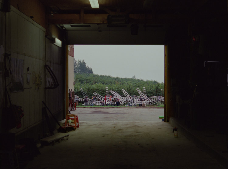 A still from Jessica Johnson and Ryan Ermacora's Labour/Leisure. Labourers on a cherry orchard carry large white ladders to a worksite, viewed from within a large warehouse door.